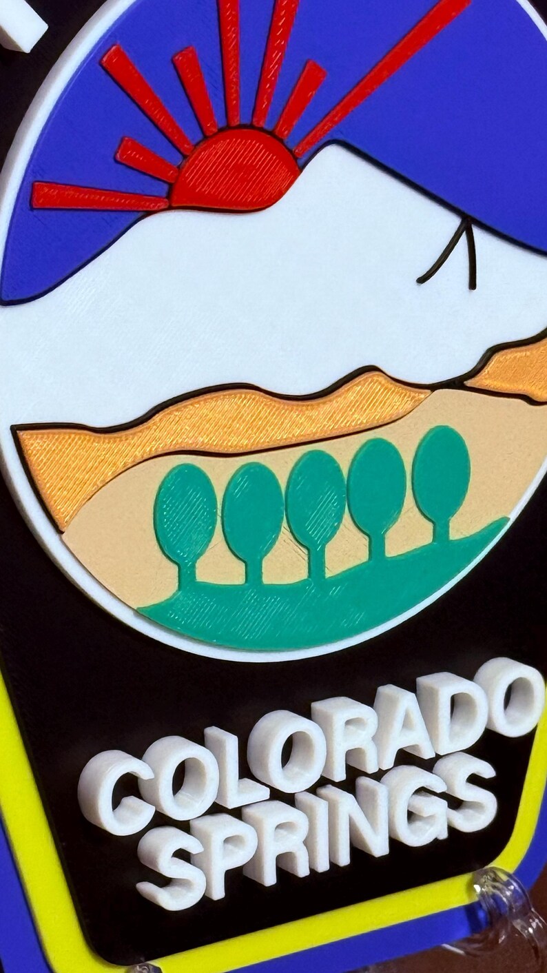 K&ouml;nnte beinhalten: Ein 3D-gedrucktes Schild mit blauem Hintergrund und einer wei&szlig;en Bergszene mit einer aufgehenden roten Sonne. Das Schild tr&auml;gt die Aufschrift "COLORADO SPRINGS" in wei&szlig;en Buchstaben.