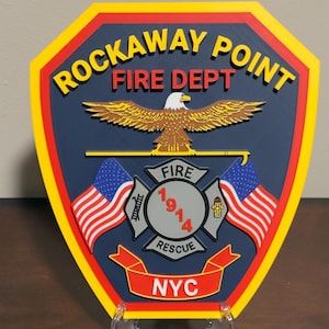 May include: A blue and yellow shield-shaped plaque with the words "Rockaway Point Fire Dept" in red and white lettering. The plaque features a bald eagle with outstretched wings, two American flags, and a fire department emblem with the words "Fire 1914 Rescue" and "NYC".