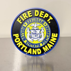 May include: A round, black and blue sign with the words "FIRE DEPT. PORTLAND MAINE" in yellow. The center features a yellow emblem with a ship, eagle, and the Latin phrase "RESURGAM".