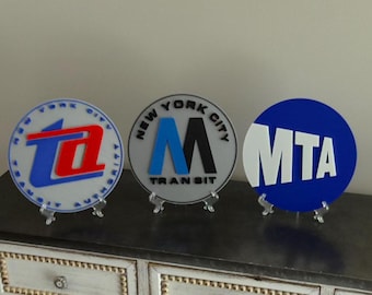 May include: Three circular signs featuring New York City transit logos. The left sign displays a red and blue TA logo with the text New York City Transit Authority. The center sign shows a blue and black M logo with New York City Transit text. The right sign features a white MTA logo on a blue background.