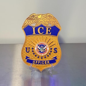 May include: A gold-colored badge with blue accents, featuring the text "ICE" at the top and "OFFICER" at the bottom. The badge has a detailed design with an eagle and the US Department of Homeland Security seal.