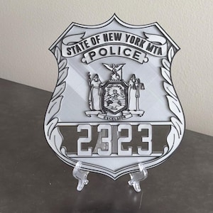 May include: A silver metal badge with the text "State of New York MTA Police" and the number "2323". The badge features a shield with the New York State seal and is on a clear plastic stand.