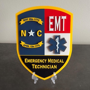 May include: A shield-shaped plaque with a yellow border, featuring the text "EMT" in white on a red background. The plaque includes a blue section with "N C" and a star, plus the text "Emergency Medical Technician".