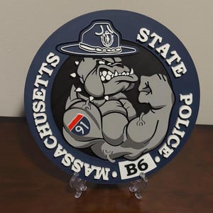 May include: A blue and white round patch with a cartoon bulldog wearing a hat that says "State Police" and "Massachusetts" around the outside. The bulldog has a number "91" on its arm.