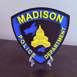 May include: A shield-shaped emblem for the Madison Police Department. The emblem features a blue border, black centre, and yellow lettering that reads "MADISON POLICE DEPARTMENT." A yellow and orange building is in the centre.