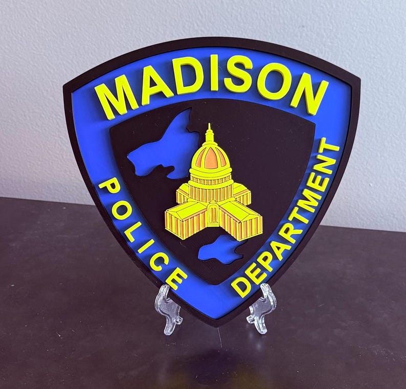 May include: A shield-shaped emblem for the Madison Police Department. The emblem features a blue border, black centre, and yellow lettering that reads "MADISON POLICE DEPARTMENT." A yellow and orange building is in the centre.