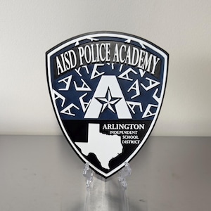 May include: A shield-shaped emblem with the text "AISD POLICE ACADEMY" in white on a black and blue background. The emblem features a white star and the outline of Texas. The text "ARLINGTON INDEPENDENT SCHOOL DISTRICT" is also visible.
