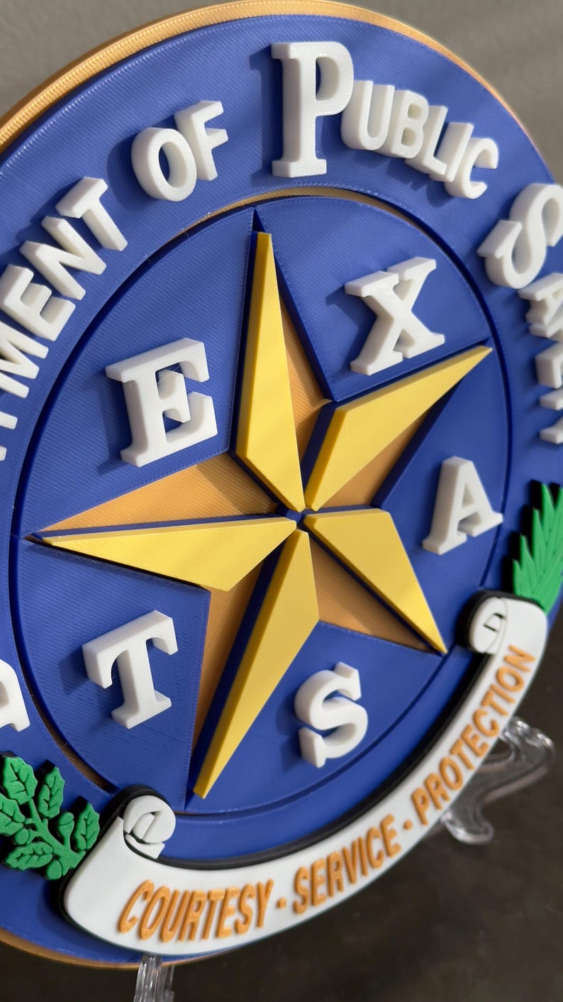 May include: A blue and gold circular sign with a five-pointed star in the center. The star is made of gold and has white letters spelling out "TEXAS" around it. The sign also has white lettering around the outside that reads "Department of Public Safety" and "Courtesy - Service - Protection".