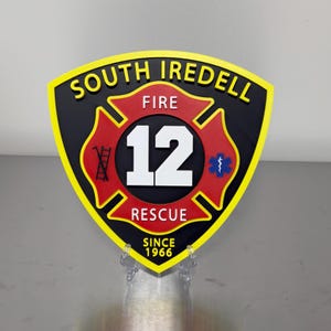 May include: A shield-shaped sign with the words "SOUTH IREDELL FIRE RESCUE" in yellow lettering. The center features a red Maltese cross with the number "12" in white. The sign also includes the text "SINCE 1966".