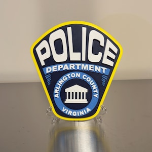 May include: A shield-shaped Arlington County Police Department sign featuring a dark blue background with white text reading POLICE DEPARTMENT ARLINGTON COUNTY VIRGINIA and a white building icon, all outlined in a bright yellow border.