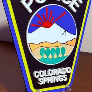 K&ouml;nnte beinhalten: Ein 3D-gedrucktes Polizeibadge mit blauem und gelbem Rand. Das Abzeichen zeigt einen wei&szlig;en Berg mit einer aufgehenden roten Sonne dahinter und den Text "POLICE" und "COLORADO SPRINGS" in Wei&szlig;.