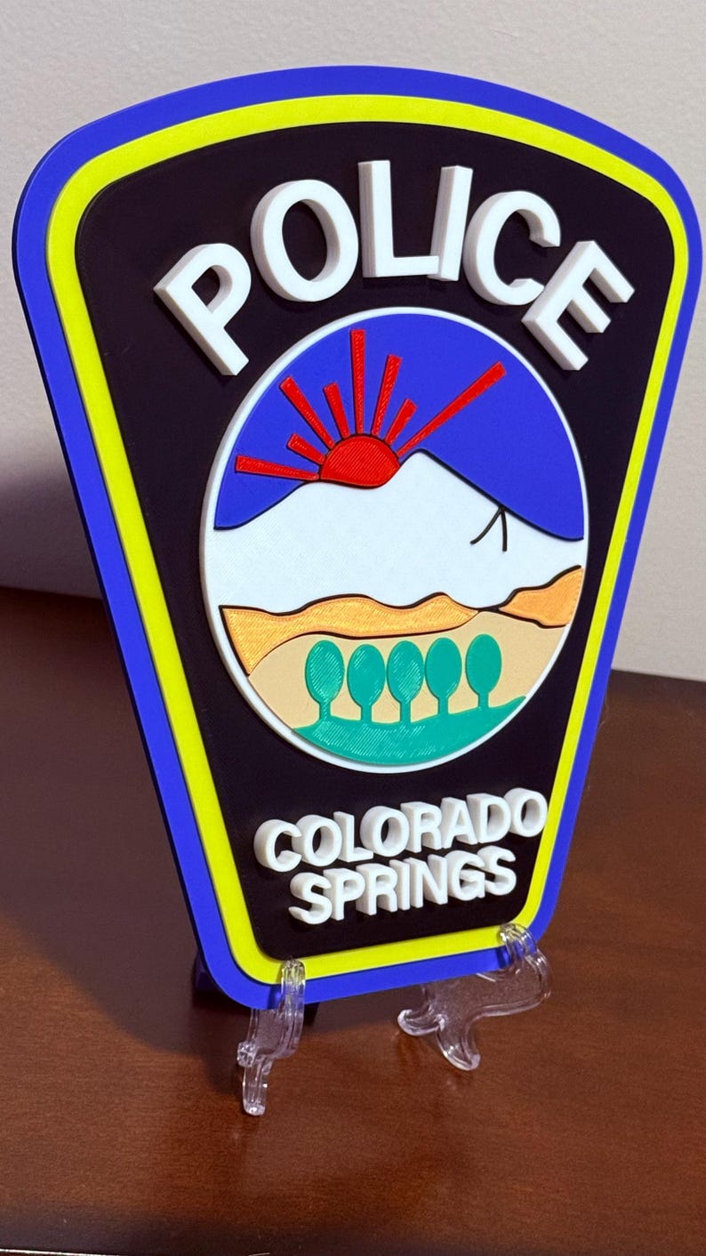 K&ouml;nnte beinhalten: Ein 3D-gedrucktes Polizeibadge mit blauem und gelbem Rand. Das Abzeichen zeigt einen wei&szlig;en Berg mit einer aufgehenden roten Sonne dahinter und den Text "POLICE" und "COLORADO SPRINGS" in Wei&szlig;.