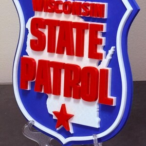 May include: A blue, white, and red Wisconsin State Patrol emblem. The shield-shaped sign features the words "WISCONSIN STATE PATROL" in red, with a white outline and a red star. The sign is mounted on a clear stand.