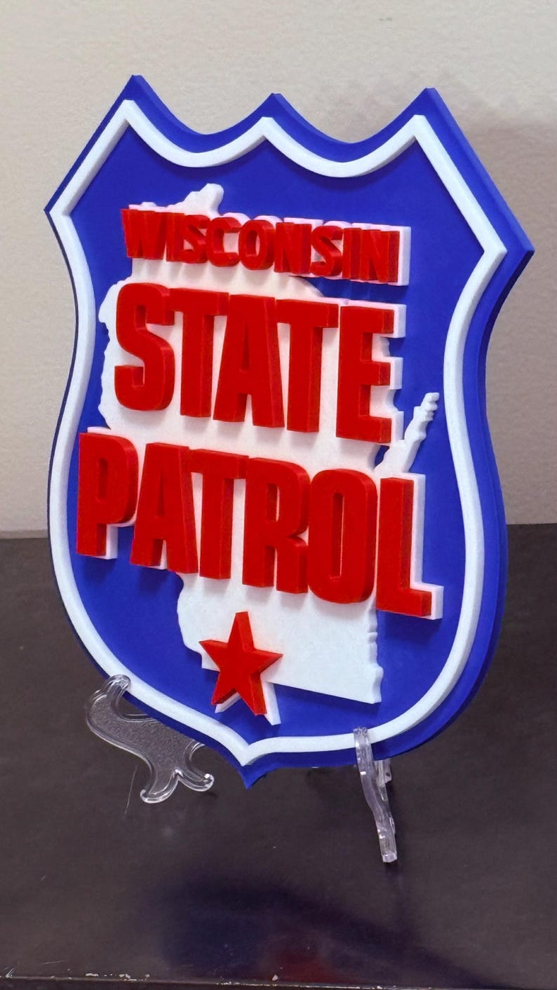 May include: A blue, white, and red Wisconsin State Patrol emblem. The shield-shaped sign features the words "WISCONSIN STATE PATROL" in red, with a white outline and a red star. The sign is mounted on a clear stand.
