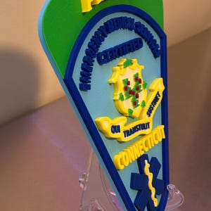 Op de afbeelding: AEMT-certificeringsplaat met een groen, blauw en geel kleurenschema. De plaat toont de tekst "AEMT", "Emergency Medical Services Certified" en "Connecticut". Het zegel van de staat Connecticut is ook afgebeeld. Transparante standaard.