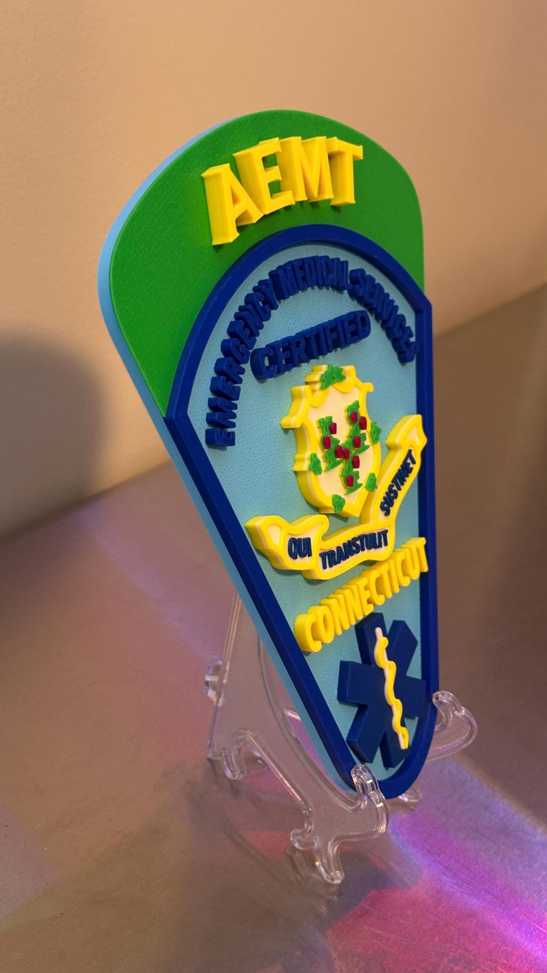 Op de afbeelding: AEMT-certificeringsplaat met een groen, blauw en geel kleurenschema. De plaat toont de tekst "AEMT", "Emergency Medical Services Certified" en "Connecticut". Het zegel van de staat Connecticut is ook afgebeeld. Transparante standaard.