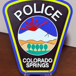 K&ouml;nnte beinhalten: Ein schwarzes und blaues Polizeibadge mit den Worten "POLICE" und "COLORADO SPRINGS" in wei&szlig;en Buchstaben. Das Abzeichen zeigt eine stilisierte Bergkette mit einer aufgehenden roten Sonne dahinter.