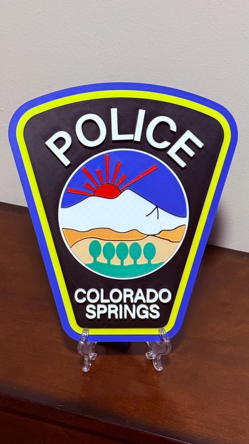 K&ouml;nnte beinhalten: Ein schwarzes und blaues Polizeibadge mit den Worten "POLICE" und "COLORADO SPRINGS" in wei&szlig;en Buchstaben. Das Abzeichen zeigt eine stilisierte Bergkette mit einer aufgehenden roten Sonne dahinter.