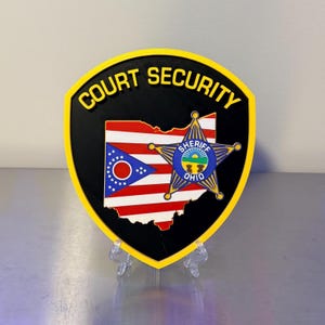 May include: A shield-shaped emblem with the words "COURT SECURITY" in yellow lettering. The emblem features the Ohio state flag and a sheriff's star. The shield is black with a yellow border and is displayed on a clear stand.