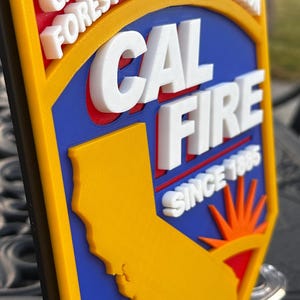 May include: A multi-layered emblem featuring the text "CAL FIRE" and "CALIFORNIA DEPARTMENT OF FORESTRY & FIRE PROTECTION". The emblem includes a yellow outline, a blue background, and a yellow silhouette of California. The text "SINCE 1885" is also visible.