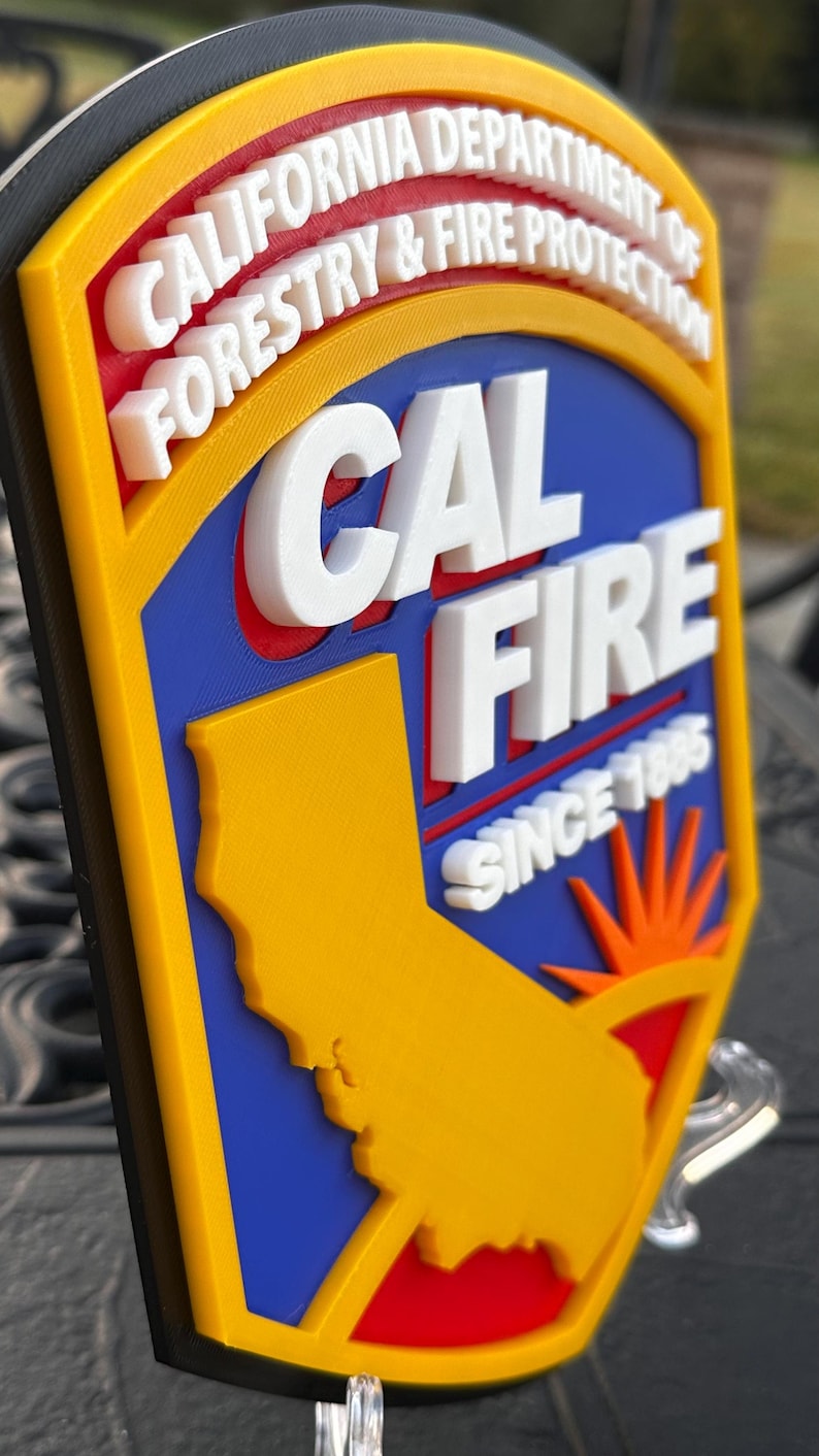 May include: A multi-layered emblem featuring the text "CAL FIRE" and "CALIFORNIA DEPARTMENT OF FORESTRY & FIRE PROTECTION". The emblem includes a yellow outline, a blue background, and a yellow silhouette of California. The text "SINCE 1885" is also visible.