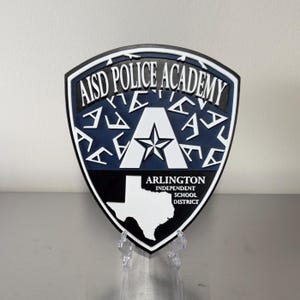 May include: A shield-shaped wall plaque featuring the text AISD POLICE ACADEMY at the top, a large letter A with a star in the center, and a white map of Texas with the text ARLINGTON INDEPENDENT SCHOOL DISTRICT in a dark blue and white color scheme.