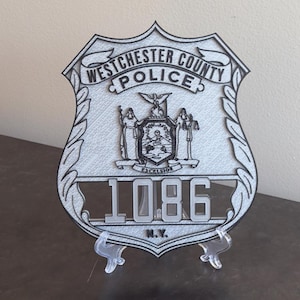May include: A silver-colored police badge with the text "Westchester County Police" at the top. The badge features a crest with the word "Excelsior" and the number "1086" below. The letters "N.Y." are at the bottom.