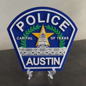 May include: A blue and white police badge with the words "Police", "Capital of Texas", and "Austin" in white lettering. The badge features a yellow star with a white building in the center, surrounded by green laurel wreaths.