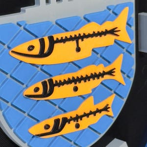 May include: A 3D-printed emblem featuring three yellow fish with black details on a blue background, set within a shield shape. The word "POLICE" is visible at the bottom. A building is at the top.