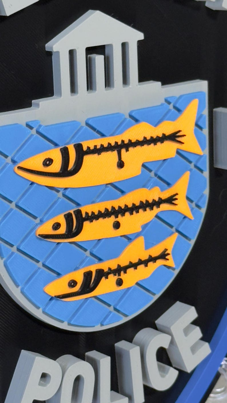 May include: A 3D-printed emblem featuring three yellow fish with black details on a blue background, set within a shield shape. The word "POLICE" is visible at the bottom. A building is at the top.