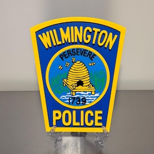 May include: A yellow and blue Wilmington Police Department emblem. The emblem features the words "WILMINGTON" and "POLICE" in yellow, with "PERSEVERE" above a yellow beehive design. The year "1739" is also displayed.