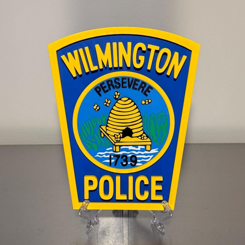 May include: A yellow and blue Wilmington Police Department emblem. The emblem features the words "WILMINGTON" and "POLICE" in yellow, with "PERSEVERE" above a yellow beehive design. The year "1739" is also displayed.