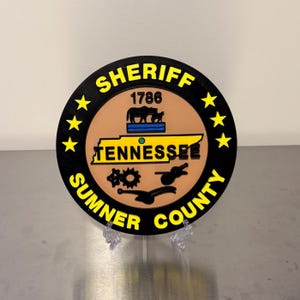 May include: A round black and yellow sign with the words "SHERIFF SUMNER COUNTY" and "TENNESSEE" in yellow. The sign has a brown center with a blue and yellow Tennessee outline and the year 1786.