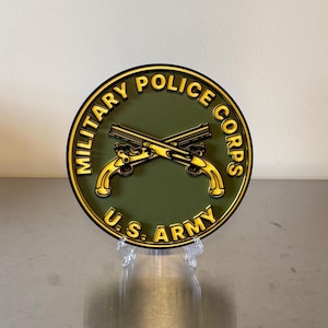 May include: A circular Military Police Corps U.S. Army emblem featuring a dark olive green background with raised yellow lettering and a central graphic of two crossed antique pistols. The emblem is displayed on a clear plastic stand.