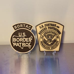 May include: Two layered wooden wall plaques. The left plaque is circular with a top arch, featuring the text BORTAC and U.S. BORDER PATROL with a map outline. The right plaque is shield-shaped, displaying U.S. CUSTOMS AND BORDER PROTECTION text.