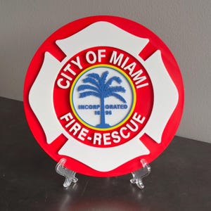 May include: A circular City of Miami Fire-Rescue emblem featuring a red background with a white Maltese cross overlay. The center displays a blue palm tree graphic with the text INCORPORATED 1896, surrounded by a yellow ring and white lettering.