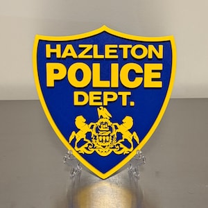 May include: A blue and yellow Hazleton Police Dept. emblem. The shield-shaped emblem features the words "HAZLETON POLICE DEPT." in yellow block letters. The emblem also includes a gold crest with two lions.