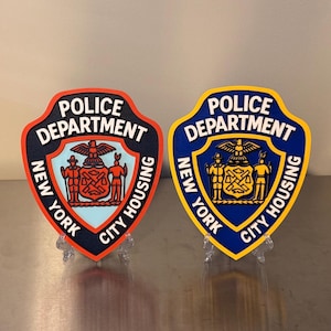 May include: Two shield-shaped wall plaques representing the New York City Housing Police Department. The left plaque features a light blue center with an orange border, while the right plaque has a blue center with a yellow border and white text.
