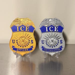 May include: Two metal badges, one gold and one silver, displayed on clear stands. The gold badge reads "ICE OFFICER" with "U.S." and a central emblem. The silver badge reads "ICE TASK FORCE OFFICER" with "U.S." and a central emblem.