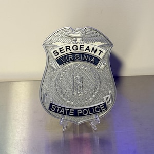 May include: A silver-toned Virginia State Police Sergeant badge featuring an eagle at the top, the text SERGEANT, VIRGINIA, and STATE POLICE in dark blue lettering, and a central circular emblem depicting a figure standing within a wreath.
