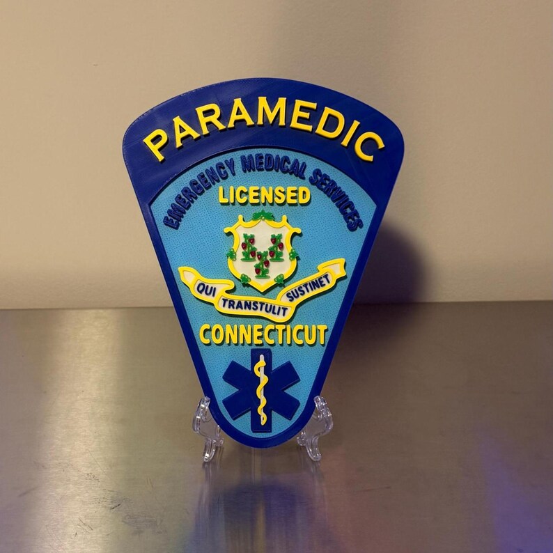 Op de afbeelding: Een blauw en geel Paramedicus-badge uit Connecticut, met de woorden "Emergency Medical Services" en "Licensed". De badge heeft een levensster-symbool en de staatsmotto "Qui Transtulit Sustinet."