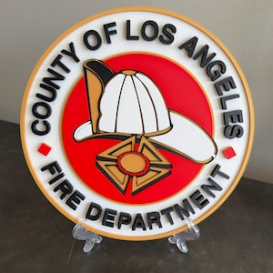 May include: A round, white plaque with a gold border and the words "County of Los Angeles Fire Department" in black lettering. The center of the plaque features a red circle with a white firefighter's helmet and a gold star with red accents.