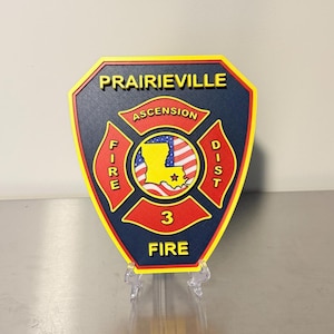 May include: A shield-shaped emblem with the text "Prairieville" in gold. The emblem features a red and gold Maltese cross design with the words "Fire" and "Dist" on either side. The center has a map of Louisiana and the text "Ascension 3 Fire".