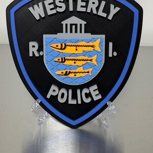 May include: A shield-shaped sign with the words "WESTERLY POLICE R.I." in white lettering. The sign has a black background with a blue border. The center features a blue square with three yellow fish.