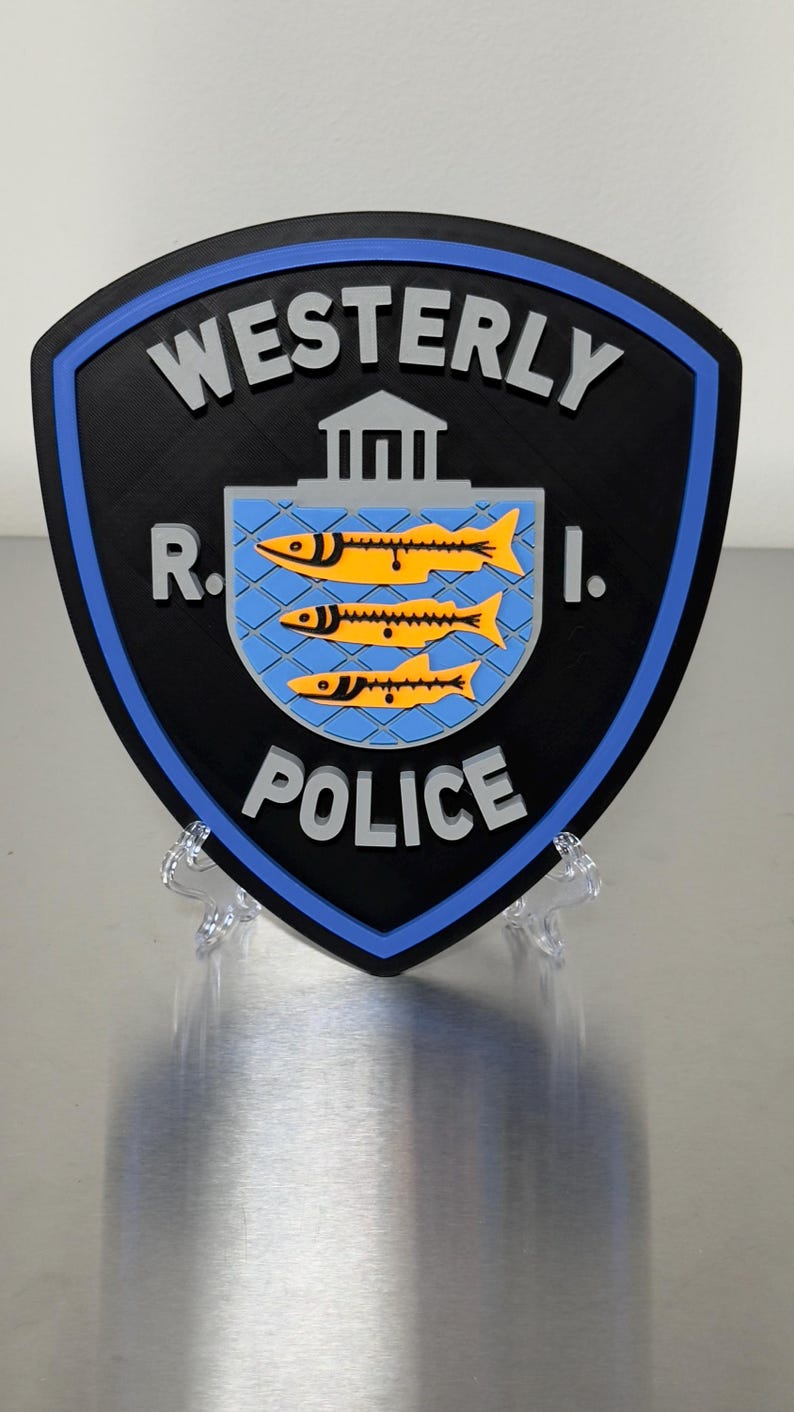 May include: A shield-shaped sign with the words "WESTERLY POLICE R.I." in white lettering. The sign has a black background with a blue border. The center features a blue square with three yellow fish.