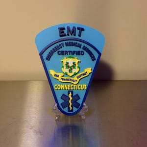 Op de afbeelding: Een blauwe EMT-badge uit Connecticut, met de woorden "Emergency Medical Services Certified". De badge bevat een staatszegel en een symbool van de levensster. De tekst "EMT" staat bovenaan.