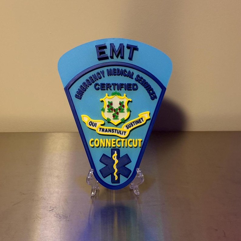 Op de afbeelding: Een blauwe EMT-badge uit Connecticut, met de woorden "Emergency Medical Services Certified". De badge bevat een staatszegel en een symbool van de levensster. De tekst "EMT" staat bovenaan.