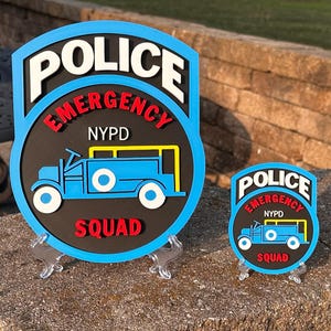 May include: Two layered signs featuring a black circular center with a blue border. The text reads POLICE in white, EMERGENCY in red, NYPD in white, and SQUAD in red. A blue truck graphic is centered on each sign. Both are displayed on clear stands.