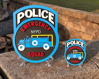 May include: Two layered signs featuring a black circular center with a blue border. The text reads POLICE in white, EMERGENCY in red, NYPD in white, and SQUAD in red. A blue truck graphic is centered on each sign. Both are displayed on clear stands.
