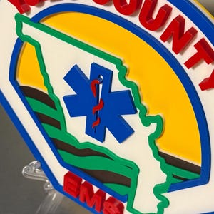 May include: A circular sign featuring the words "YOLO COUNTY" in red, with a blue border. The sign includes a blue star of life symbol and a green outline of a state. The letters "EMS" are in red.
